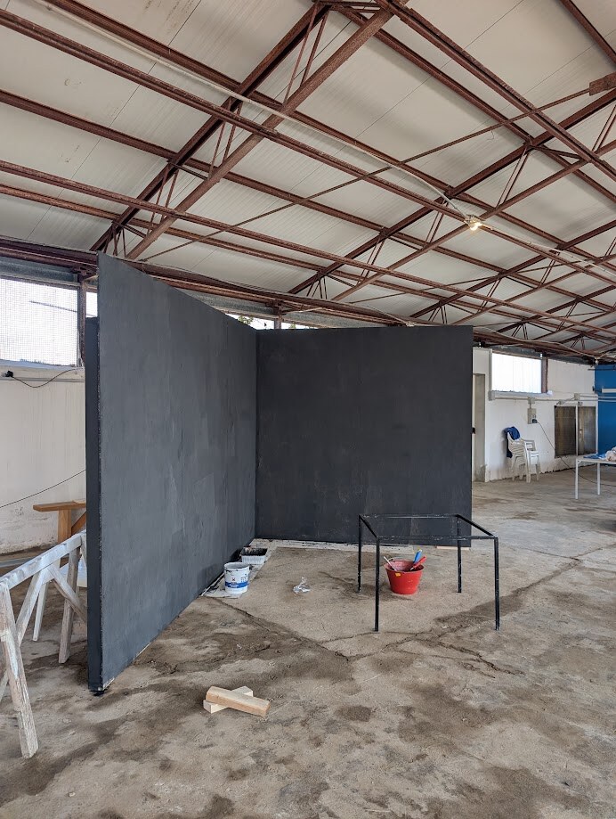 An industrial shed with metal trusses; two matte-black walls being erected in an L-shape, a small work table with paint buckets, sawhorses to the side. The room being prepared for the installation.