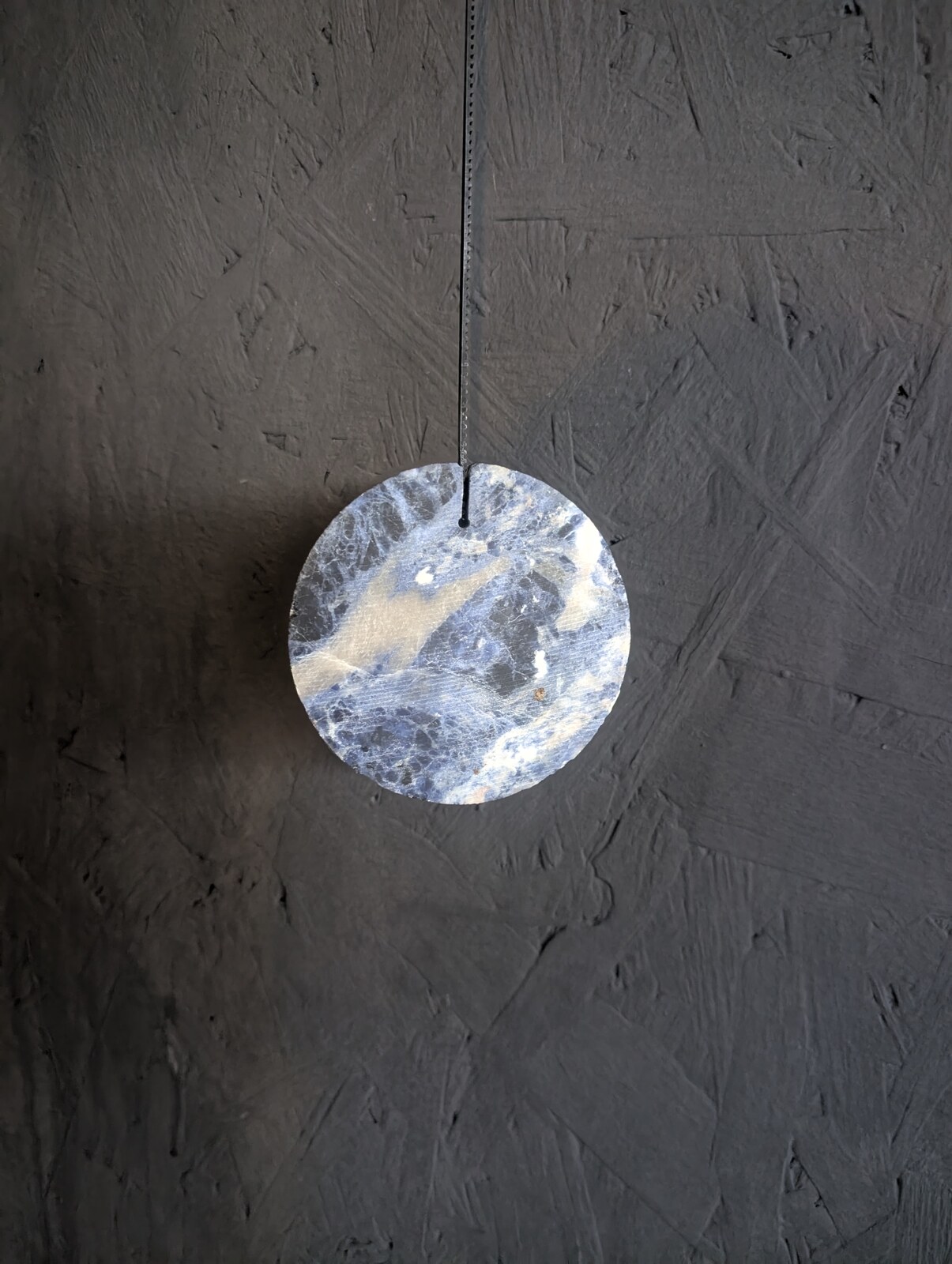 A pale marbled wooden disc hanging from a thin cable against a dark textured plaster wall.