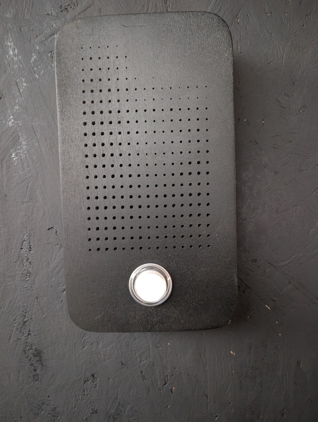 A matte-black plate mounted on grey plaster, its surface perforated in a dense dot matrix. A small round diffuser glows warm-white near the lower edge.