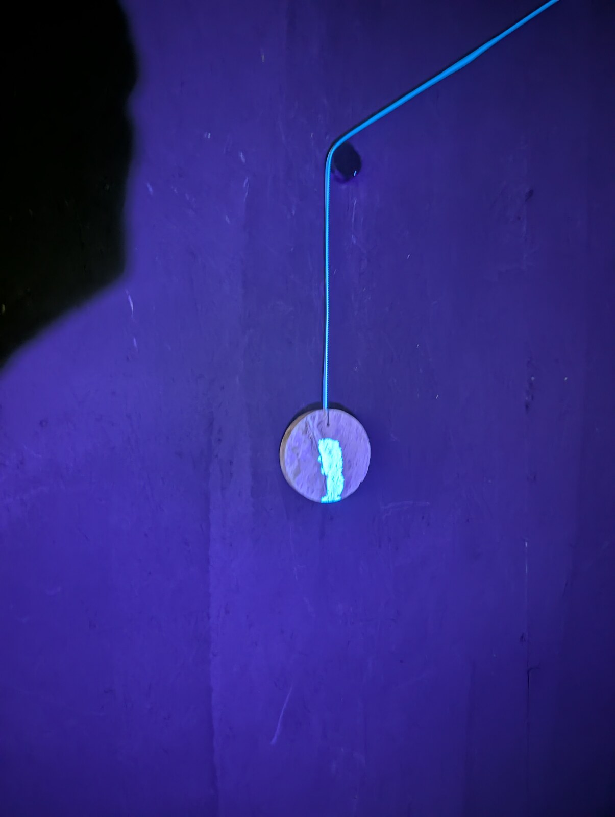 A single wooden disc suspended from a bright cable against a saturated ultraviolet wall; a glowing pale mark on its surface.