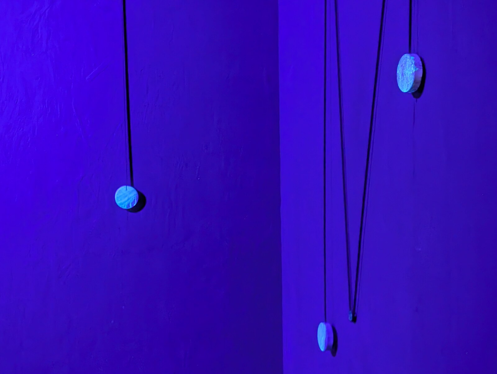 Three wooden discs hanging at different heights on dark cables in a corner lit by ultraviolet light; the walls read deep violet-blue.