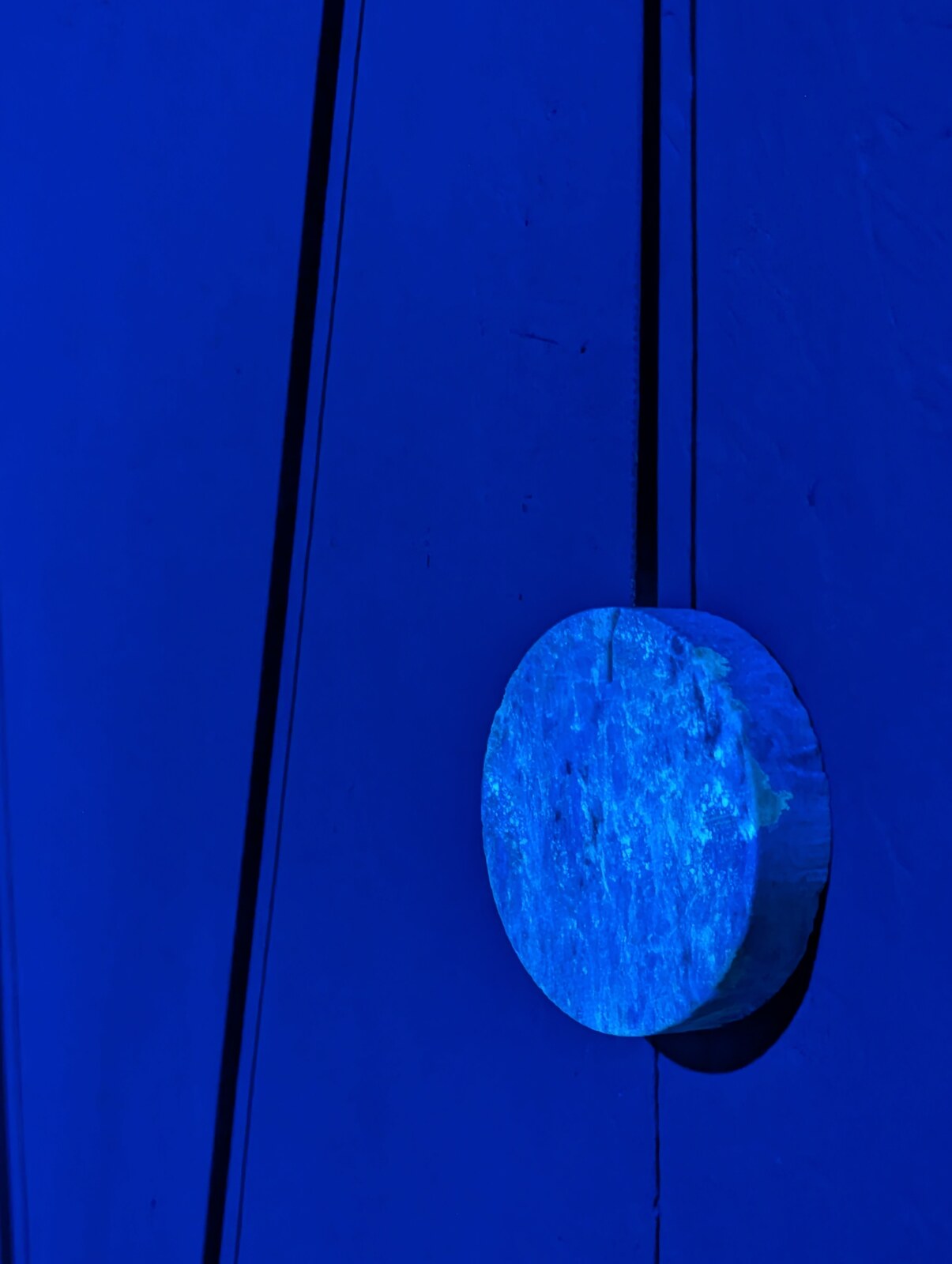 Close view of a wooden disc, painted cobalt-blue, resting flat against a saturated cobalt-blue wooden surface; the disc casts a soft shadow.
