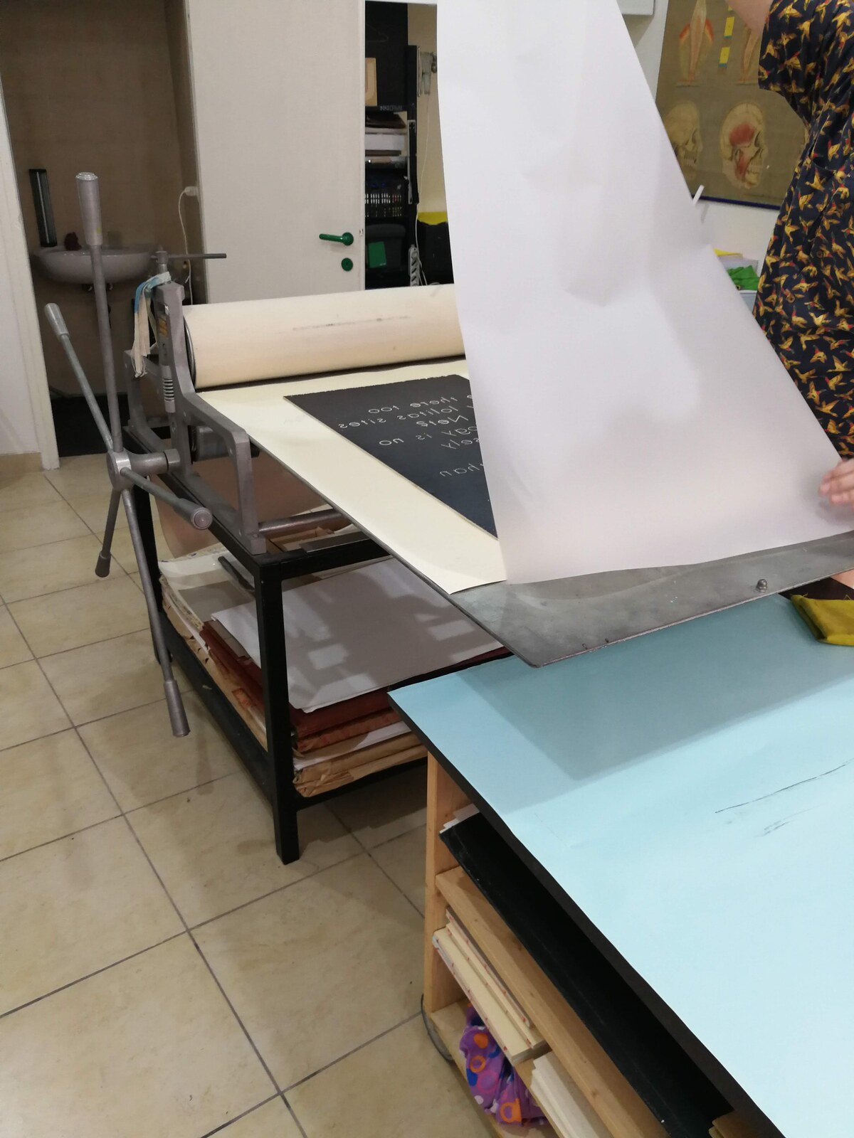 A calcographic press in a studio; a black-and-white poster is being lifted from the press bed, revealing fresh print. An anatomical human-body poster hangs on the wall in the background.