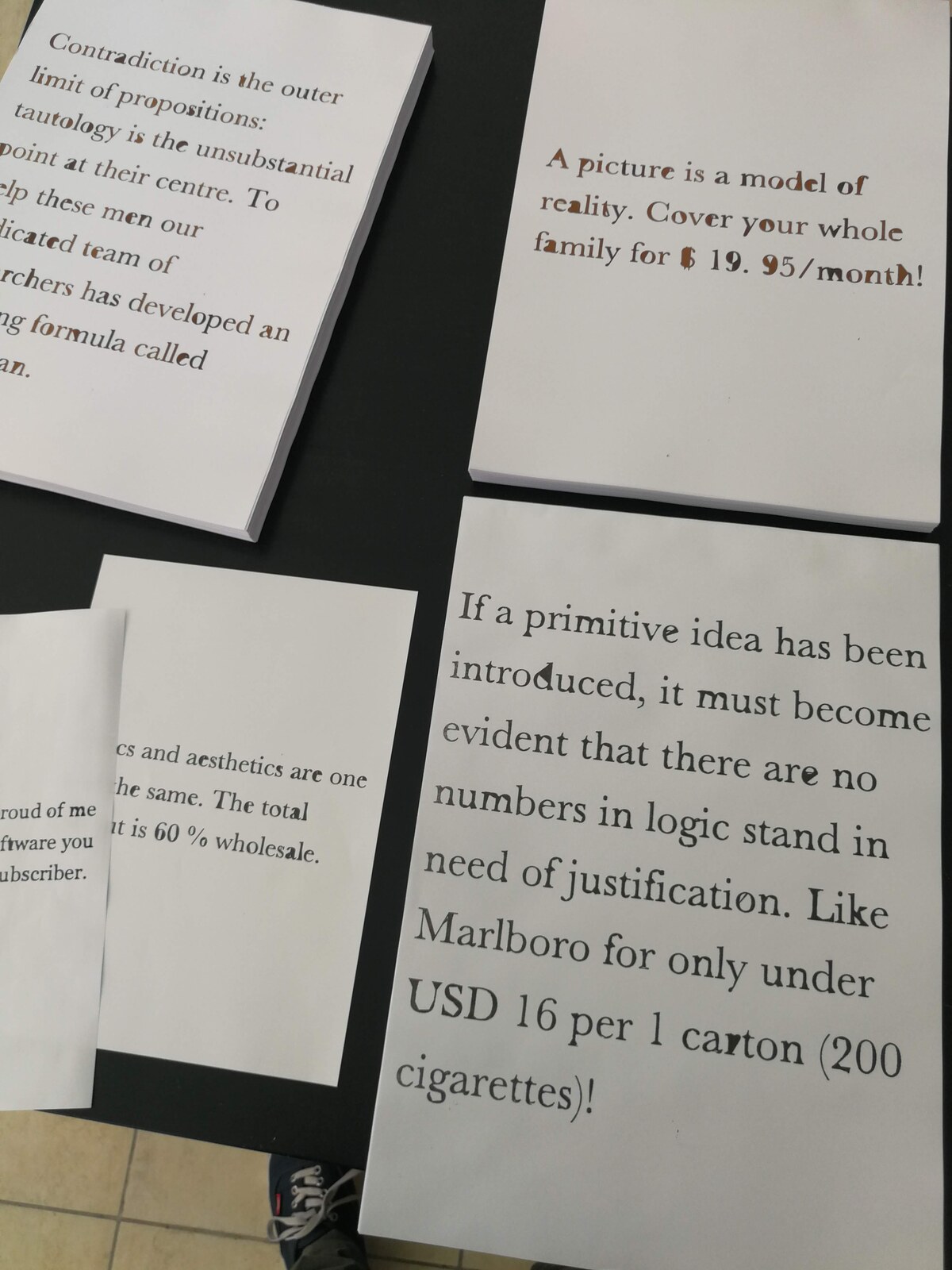 Four small printed booklets on a black table, each carrying a text that collages a proposition from Wittgenstein's Tractatus with a fragment of spam email — one visible line reads 'A picture is a model of reality. Cover your whole family for $19.95/month!'