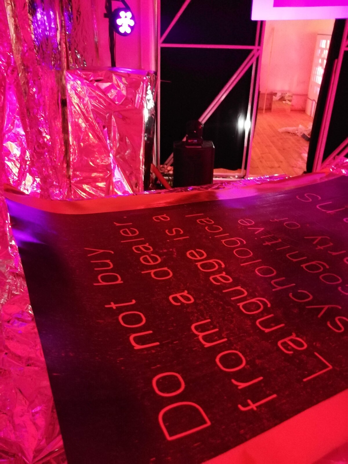 Night view inside the None collective's space in Rome: crumpled silver-and-pink tinfoil sheets on floor and wall, a large red-on-black poster in the foreground reading 'Do not buy language from a dealer'.