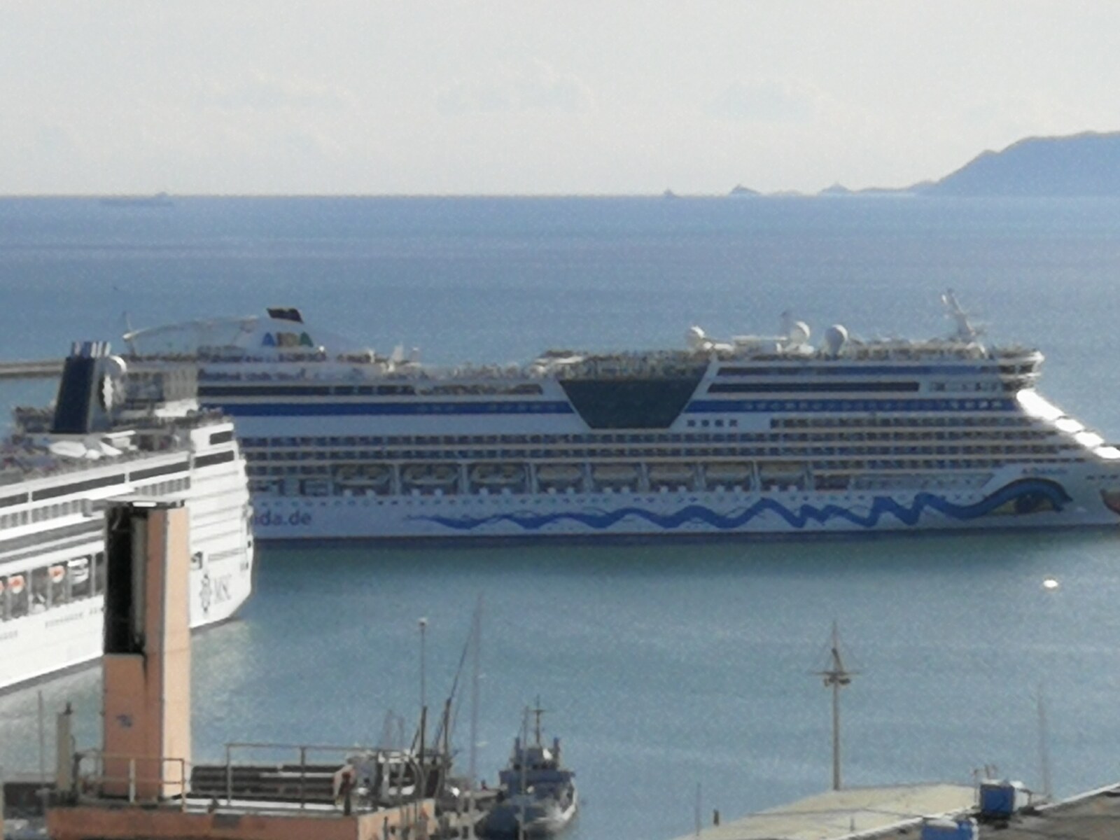 An AIDA cruise ship dwarfing a smaller MSC ferry, docked in a Mediterranean port on a hazy afternoon.