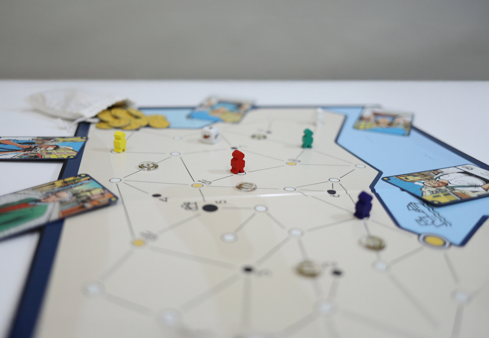 Close view of the UNSEASONED boardgame in play: a hexagonal network of nodes, wooden pieces in yellow, red, green and purple, a scattered pile of gold coins, illustrated cards along the edges.