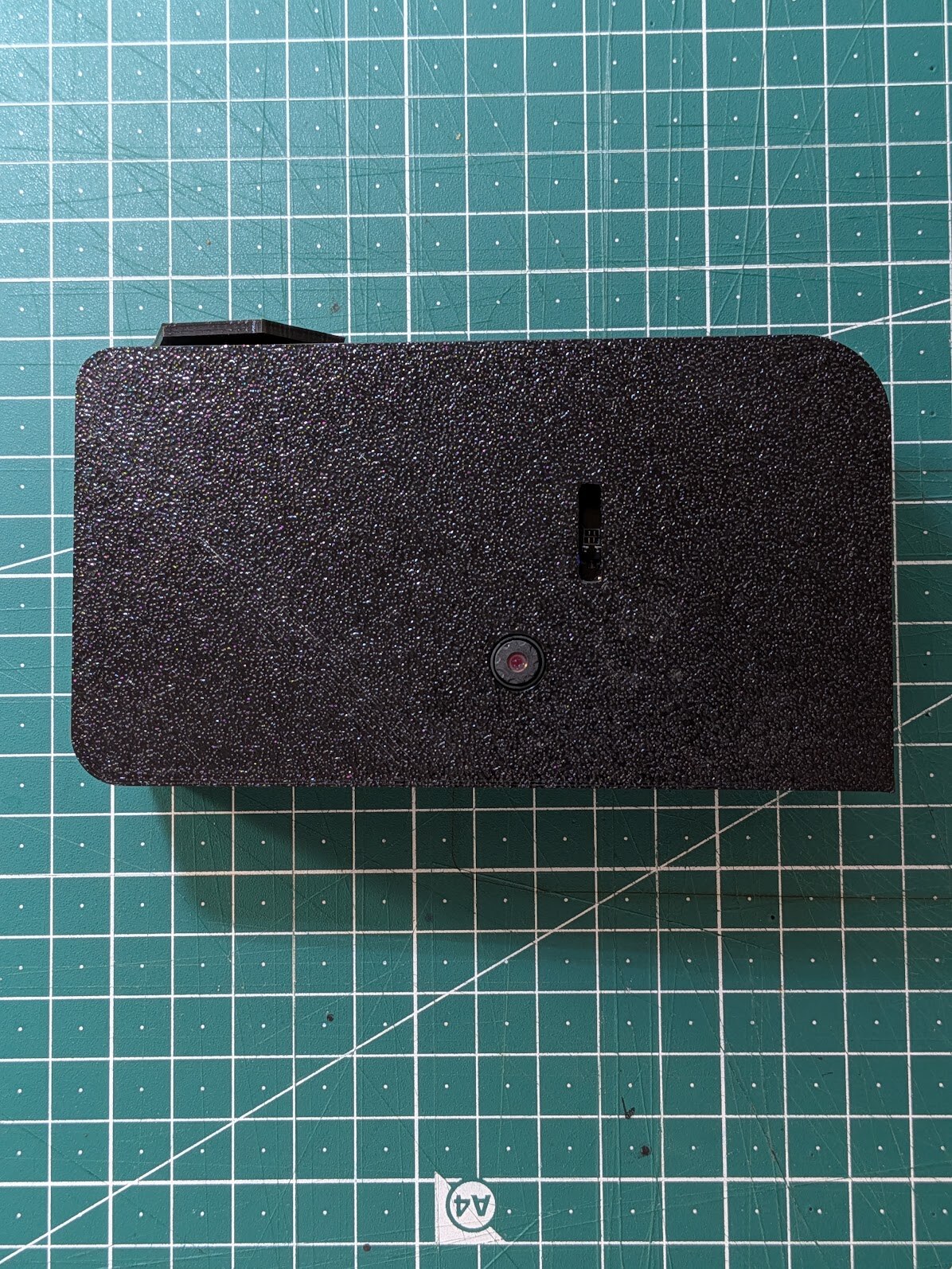 A matte black 3D-printed camera body lying face-up on a green cutting mat. A slide switch and one small lens are the only openings in an otherwise blank slab.