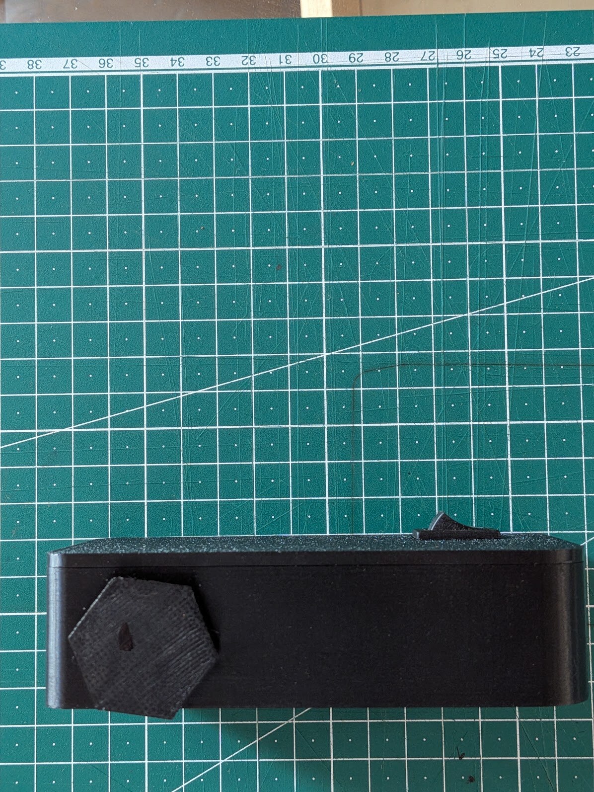 The same body seen from its edge: a hexagonal printed selector dial protrudes from one end, a small toggle sits on the top face. Millimetre grid of the cutting mat behind it.